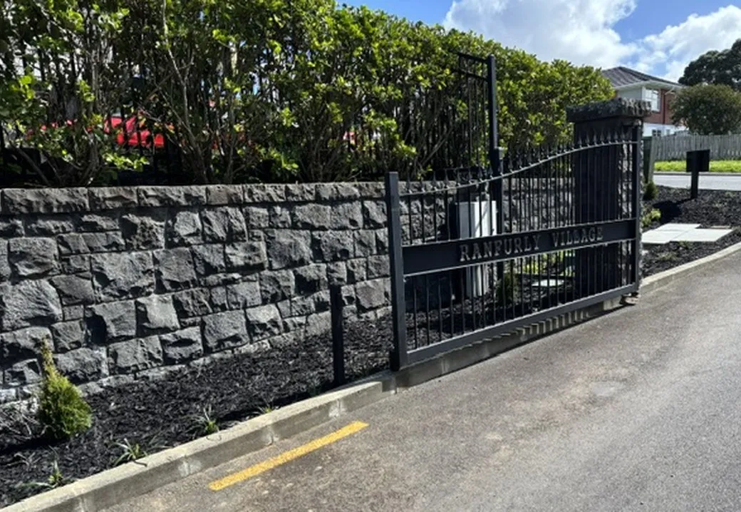 A stone boundary wall and freshly mulched bed running up to a gated village entrance