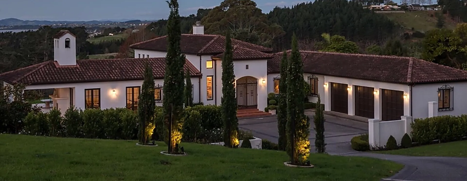 A Spanish-style Auckland home at dusk, with clipped hedging, uplit cypresses and lawn running down to the drive